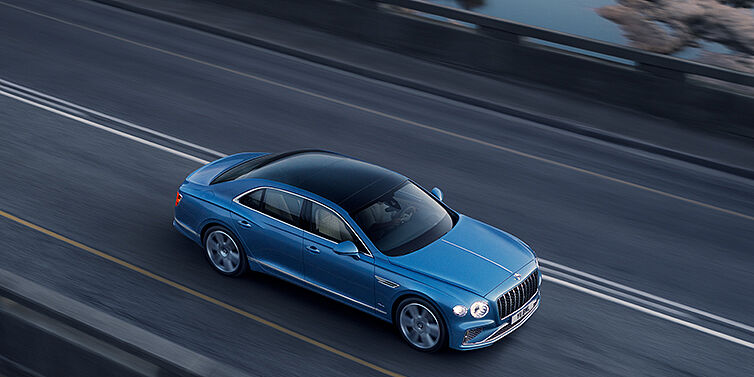 Bentley Riga New Bentley Flying Spur Azure sedan in Light windsor blue paint by Mulliner, with 22 inch Azure wheels seen from overhead driving dynamically on a country road