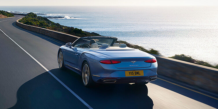 Bentley Riga New Bentley Continental GTC Azure convertible rear view in Light Windsor blue paint with 22 inch ten spoke wheels driving dynamically on an ocean road in the sunshine